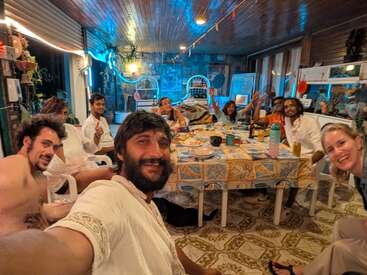 A group of friends sit around a table, smiling and waving. The room is warmly lit with blue accents, creating a festive, cozy, and welcoming atmosphere.