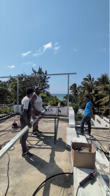 Three men work together on a rooftop under a clear blue sky, assembling metal structures. Palm trees and the ocean form a scenic, tropical background.