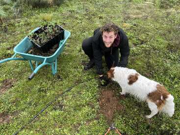 Un hombre está plantando plantones al aire libre, sonriendo a la cámara. Un perro blanco y marrón está cerca. Hay una carretilla verde llena de plantas y tierra.