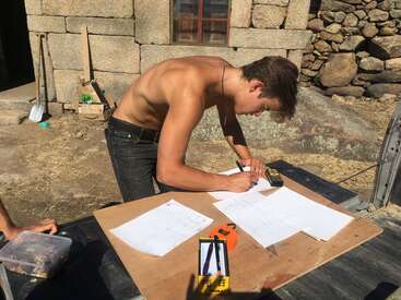 Un hombre sin camiseta se inclina sobre una mesa al aire libre, revisando planos y tomando medidas. Hay herramientas de construcción y papeles esparcidos, con muros de piedra al fondo.