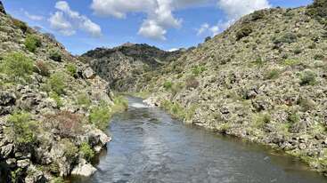 A serene river flows between rocky, green hills under a bright blue sky filled with fluffy white clouds, creating a peaceful and picturesque natural landscape.