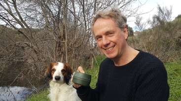 A smiling man sits outdoors holding a cup, accompanied by a white and brown dog. They are surrounded by trees and grass near a quiet river.