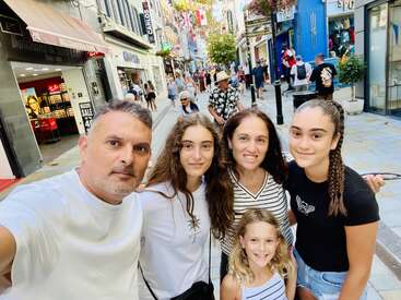 Una familia de cinco miembros, con una sonrisa en los labios, posa para hacerse un selfi en una animada y bulliciosa calle comercial, con escaparates llenos de colorido, banderas y otras personas que disfrutan de su día al aire libre.