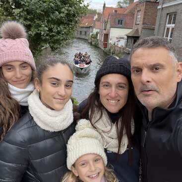 A happy family of five, bundled in winter clothes, poses for a selfie by a picturesque canal with a boat full of tourists and charming houses behind.
