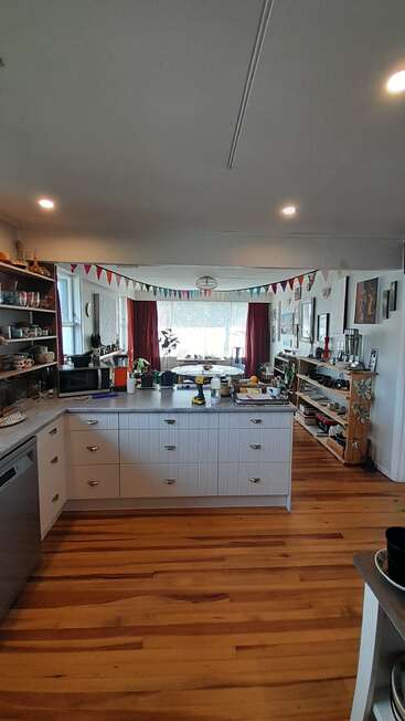 A cozy, lived-in kitchen with wooden floors, festive bunting, visible shelves, houseplants, sunlight streaming through windows, and a dining area in the background. Inviting atmosphere.