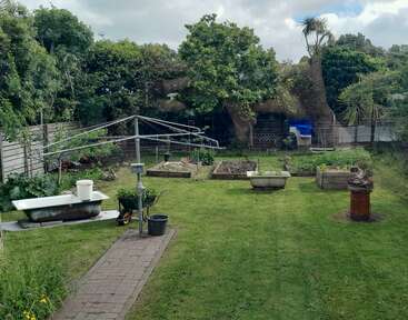 A backyard garden with raised beds, a rotary clothesline, bathtubs used as planters, lush green grass, large trees, a fence, and various gardening tools.