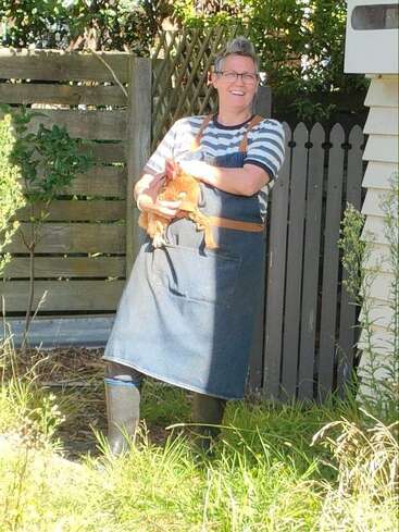 Da steht jemand lächelnd draußen, mit Brille, gestreiftem Shirt, Jeansschürze und Stiefeln, und hält fröhlich ein Huhn in einem sonnigen Gartenbereich.