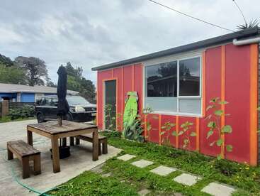 A red building with orange trim has sunflowers growing beside it. There’s a wooden picnic table, benches, and a black umbrella in the foreground. Overcast sky.
