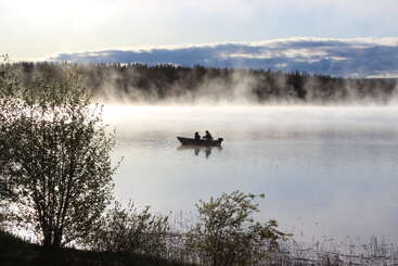 Duas pessoas estão sentadas em um barco num lago calmo e enevoado ao amanhecer. Árvores e arbustos emolduram a cena, com a floresta e as nuvens ao fundo.