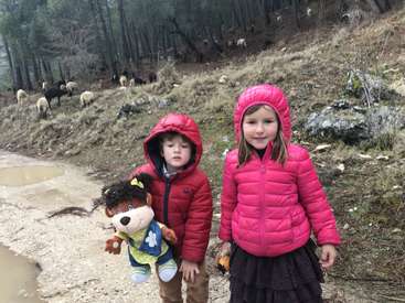 Dos niños, uno con un peluche en la mano, de pie en un camino embarrado frente a una colina con ovejas pastando al fondo, llevan abrigos de invierno.