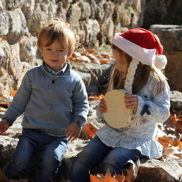 La imagen muestra a dos niños sentados sobre rocas, el de la derecha con un gorro de Papá Noel y una pandereta en la mano, mientras el otro niño mira.