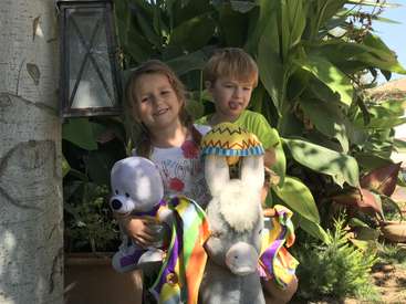 La imagen muestra a dos niños de pie, cada uno con un peluche en la mano, con exuberantes plantas verdes y una farola como telón de fondo.