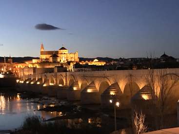 La imagen representa una serena escena de un puente que cruza un río, con un majestuoso edificio al fondo, bañado por una cálida luz, sobre un cielo azul despejado.
