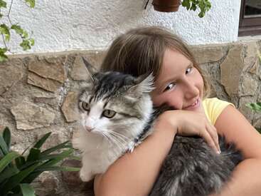 La imagen muestra a una joven acunando a un gran gato, con la cabeza del felino apoyada en su hombro, sobre el fondo de un muro de piedra.