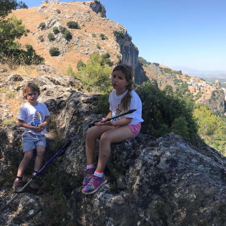 La imagen muestra a dos niños sentados en una roca, uno con un palo en la mano y el otro con una camiseta blanca y pantalones cortos.