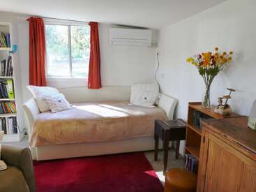 A cozy, sunlit room with a white daybed, pillows, red curtains, air conditioner, bookshelf, wooden furniture, red rug, and a vase of colorful flowers.