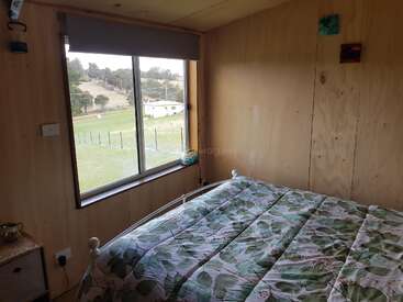 This cozy bedroom features wooden walls, a bed with leaf-patterned bedding, a large window with a scenic view, minimal decor, and natural light streaming in.