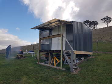 A small, elevated metal cabin stands on grassy terrain. A person tends a campfire nearby, with benches and trees in the background under a cloudy sky.