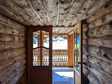 Das Bild zeigt das Innere einer rustikalen Blockhütte mit einer Glastür, die auf eine Veranda führt, mit aufwendigen Holzarbeiten und einem schönen Blick nach draußen.