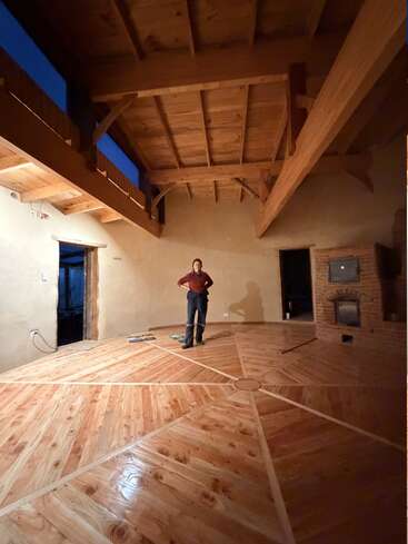 Eine Person steht in der Mitte eines geräumigen Raums mit hohen Holzdecken, einem schönen geometrischen Holzboden, rustikalen Wänden und einem gemauerten Kamin. Warme Beleuchtung.