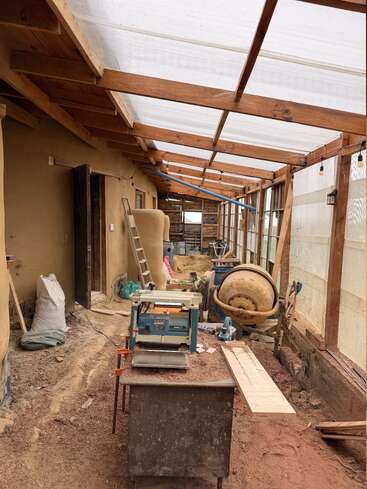 Dieses Bild zeigt eine teilweise überdachte Baustelle mit Holzbalken, Werkzeugen, Sägemehl, einer Werkbank, einem Zementmischer, einer Leiter und unfertigen Lehmmauern, was auf laufende Bauarbeiten hindeutet.