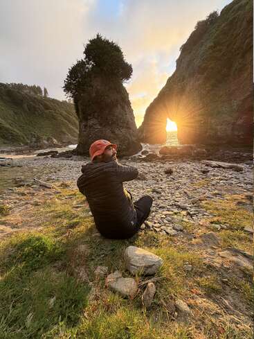 Eine Person mit einem orangefarbenen Hut sitzt auf felsigem Boden und blickt auf einen atemberaubenden Sonnenuntergang, der durch einen natürlichen Torbogen am Meer, umgeben von Klippen, scheint.