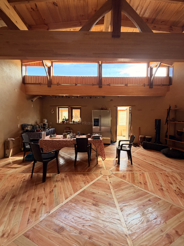 Una habitación cálida con techos y suelos de madera, grandes ventanas, una mesa de comedor con sillas, electrodomésticos de cocina y luz natural que crean un ambiente acogedor y rústico.