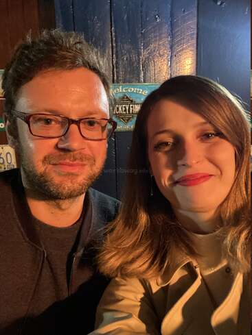 L'image montre un homme et une femme posant pour un selfie devant un mur bleu foncé sur lequel on peut lire "Welcome" et "Hockey Final".