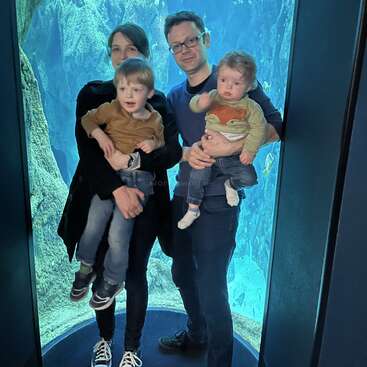 A family of four stands inside an aquarium tunnel, with blue water and fish in the background. The parents each hold a young child, smiling.