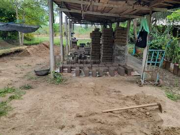 The image depicts a covered outdoor area with a dirt floor, featuring a pile of bricks, a shovel, and a cart, surrounded by trees and greenery.