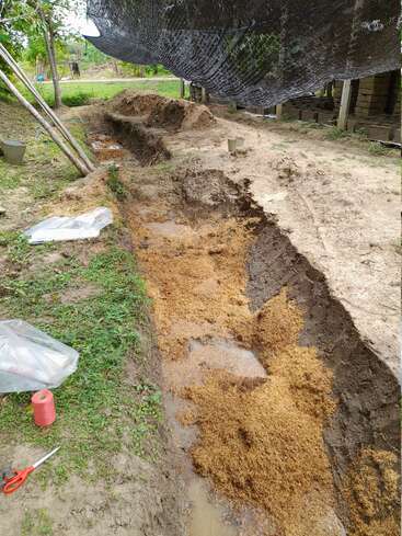The image depicts a yard with a large trench dug into the dirt, surrounded by various items such as tools, plastic bags, and a black shade cloth.