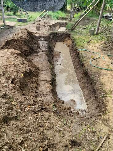 The image depicts a muddy, narrow trench with water at its bottom, surrounded by dirt and grass, with a hose and other objects nearby.