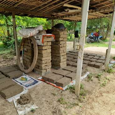 The image depicts a covered outdoor area with a pile of bricks and a motorcycle, surrounded by trees and a dirt ground, evoking a rural or construction setting.