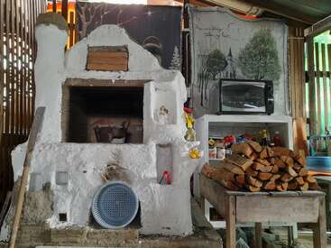 The image depicts a rustic outdoor kitchen area, featuring a white stone oven, a woodpile, and a microwave, with a serene forest scene painted on the wall.