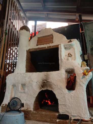 The image depicts a white, clay structure with a fireplace, featuring a small fire burning inside, and various objects on the exterior, set against a wooden background.