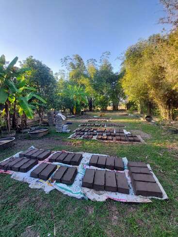 The image depicts a serene outdoor setting with a grassy area, trees, and a clear blue sky, featuring a collection of dark brown bricks arranged on a white tarp.