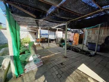 The image depicts a covered outdoor area with a brick floor, featuring a black shade cloth roof and a variety of items, including plants and bags of soil.