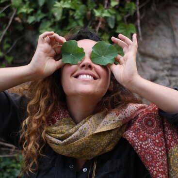 Uma mulher com longos cabelos castanhos segura duas folhas verdes sobre os olhos, em frente a um muro de pedra coberto de trepadeiras verdes. Ela usa um cachecol e uma camisa preta.