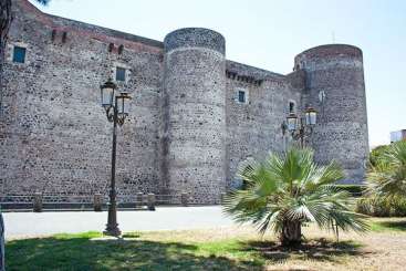 A imagem mostra um grande edifício de pedra com uma área gramada na frente, com uma palmeira e um poste de luz, em um céu azul claro.
