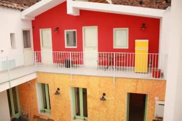 A imagem mostra um edifício de dois andares com uma varanda e um esquema de cores vibrantes, com tons de vermelho, amarelo e branco.