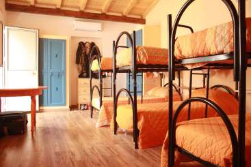 A imagem mostra um quarto com beliches, uma mesa e armários, apresentando um esquema de cores quentes com roupa de cama laranja e armários azuis, evocando uma atmosfera aconchegante de albergue ou dormitório.