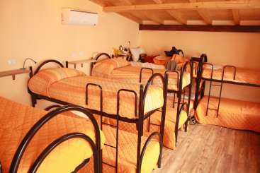 A imagem mostra um quarto com beliches, com roupa de cama laranja e armações de metal preto, situado em um piso de madeira com teto inclinado e unidade de ar-condicionado.