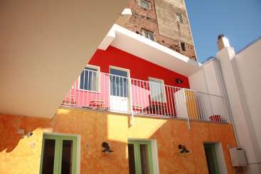 A imagem mostra um edifício vibrante de vários andares com uma varanda e uma parede vermelha, em um céu azul claro.