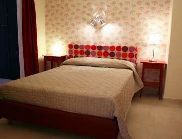 A imagem mostra um quarto com uma cama de casal, com uma cabeceira de bolinhas vermelhas e roxas, um edredom bege e uma mesa de cabeceira vermelha com uma lâmpada.