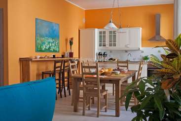 A imagem mostra uma cozinha laranja vibrante e uma área de jantar, com uma mesa de madeira, cadeiras e uma ilha de cozinha, com um sofá azul em primeiro plano.