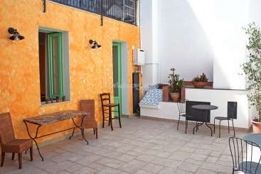 A imagem retrata um pátio externo sereno, com uma parede laranja vibrante, uma pequena mesa e cadeiras e alguns vasos de plantas, exalando uma sensação de tranquilidade.