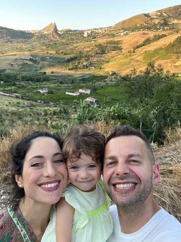 A smiling family of three poses for a selfie outdoors, with beautiful rolling hills, scattered houses, and green valleys in the scenic background under clear skies.