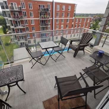 Un balcon moderne avec une balustrade en verre est équipé de plusieurs chaises et petites tables et donne sur des immeubles en briques, une route et des arbres verdoyants dans un quartier résidentiel.