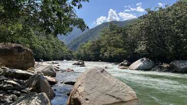 Un río cristalino discurre entre grandes rocas y frondosos árboles, con las montañas al fondo bajo un cielo azul brillante salpicado de nubes blancas y esponjosas.