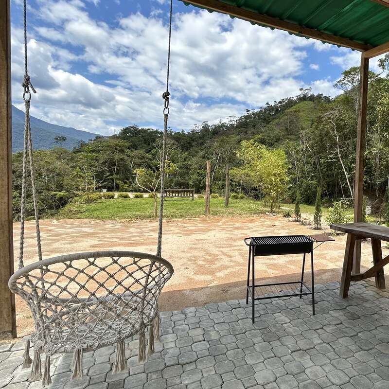 La imagen representa un entorno sereno al aire libre, con una mesa y sillas de madera, rodeado de exuberante vegetación, con un cielo despejado y una masa de agua al fondo, que evoca una sensación de tranquilidad.
